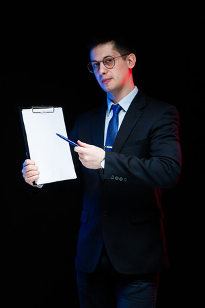 Young caucasian business man in black suit standing in office looking in camera showing on clipboard with pen thoughtful handsome confident successful sophisticated classic smart elegant modern serious isolated.の写真素材