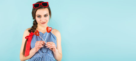 Portrait of attractive young pin-up woman in striped shirt with isolated on blue background with copyspace holding two heart-shaped candies on sticks.の写真素材