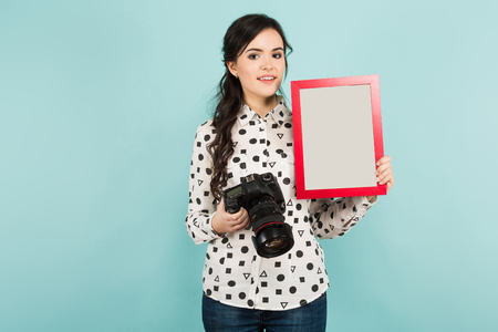 Portrait of young attractive woman photographer in white shirt holding camera and presenting a blank frame isolated on blue background with copyspace award certificate concept.の写真素材
