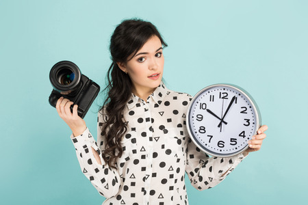 Portrait of young attractive woman photographer in white shirt holding camera and watches isolated on blue background with copyspace punctuality time is money concept.の写真素材