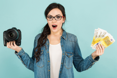 Portrait of young attractive happy woman photographer in denim and glasses shirt holding camera and fan of euro banknotes isolated on blue background with copyspace first salary or payment concept.の写真素材