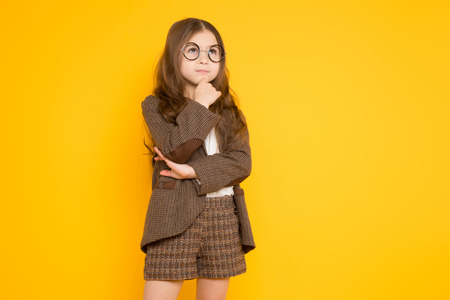 Portrait of adorable long-haired little girl in eyeglasses wearing brown jacket, shorts and white knee-socks props up her chin isolated on orange background with copyspace kids fashion concept.の写真素材