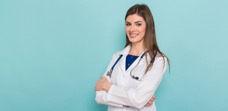 Portrait of female smiling brunette doctor with braces on her teeth in white coat with stethoscope and arms crossed isolated on blue background with copyspace head physician concept.の写真素材