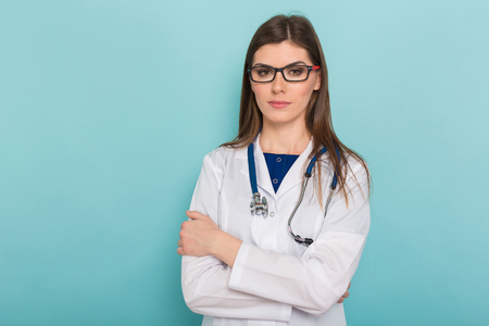 Portrait of female brunette doctor in white coat and glasses with stethoscope and arms crossed isolated on blue background with copyspace head physician concept.の写真素材
