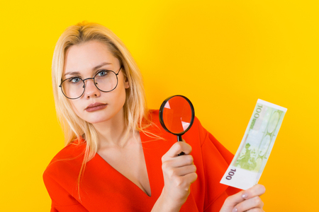 Portrait of attractive blonde woman in red dress and eyeglasses isolated on yellow background looking through magnifying glass on euro banknote checking if it is fake counterfeiter concept.の写真素材