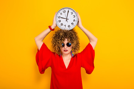 Portrait of a white woman with afrro curly hairstyle in red dress and sunglasses holding watches above her head isolated on orange background with copyspace punctuality being on time or late concept.の写真素材