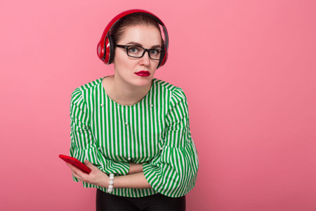 Portrait of businesswoman with hair bun in striped blouse and eyeglasses listens to music in headphones holds cellphone with arms crossed isolated on pink background with copyspace.の写真素材