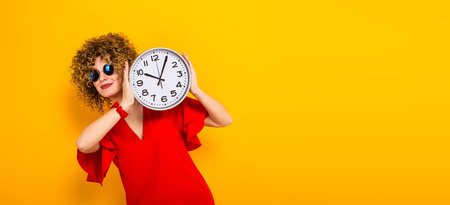 Portrait of a white woman with afrro curly hairstyle in red dress and sunglasses hiding behind watches isolated on orange background with copyspace punctuality being on time or late concept horizontal.の写真素材