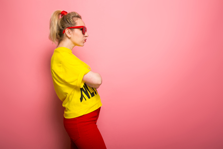 Attractive woman in yellow T-shirt, red jeans and sunglasses standing sideways with arms crossed isolated on pink background with copyspace.の写真素材