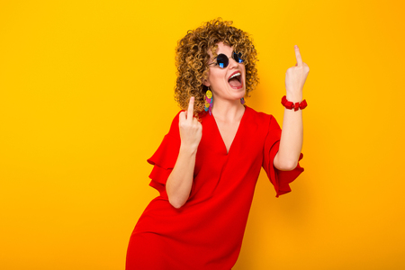 Portrait of a white woman with afrro curly hairstyle in red dress and sunglasses laughing and showing fuck sign isolated on orange background with copyspace beauty salon advertisement concept.の写真素材