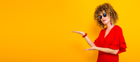 Portrait of a white woman with Afro curly hairstyle in red dress and sunglasses pointing with her hands at empty space with your text isolated on orange backgroundの写真素材