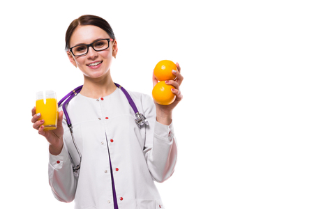 Portrait of young attractive caucasian brunette positive friendly smiling woman nutritionist standing in office with fruit mandarin orange and glass of fresh juice in her hands looking in camera in white uniform.の写真素材