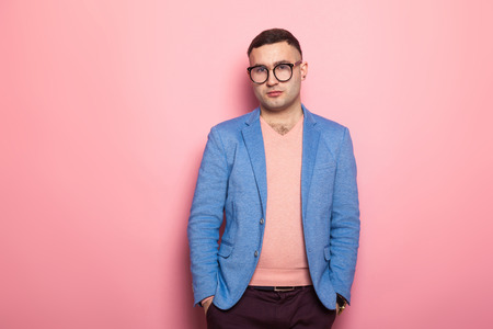 Portrait of handsome man in eyeglasses, pink jumper and blue jacket holding hands in pockets isolated on pink background with copyspace male fashion advertising concept.の写真素材