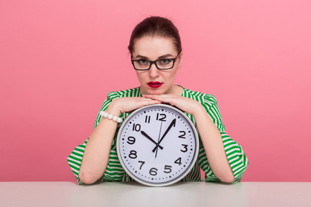 Portrait of attractive businesswoman with hair bun in striped blouse and eyeglasses showing watches sits at table isolated on pink background with copyspace punctuality being late concept.の写真素材