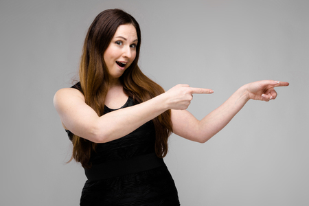 Emotional plus size model standing in studio pointing fingers outstretched hands to the right showing on blank area