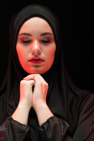 Attractive confident serious muslim arabian woman dressed in black hijab standing in studio looking in camera with hands near her face as religion conceptの写真素材