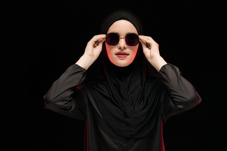Attractive confident muslim arabian woman model dressed in black hijab and sunglasses standing in studio looking in camera with hand on hand as fashion conceptの写真素材