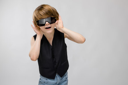 Portrait of cute little boy wearing black vest and stylish sunglasses making faces isolated on grey...