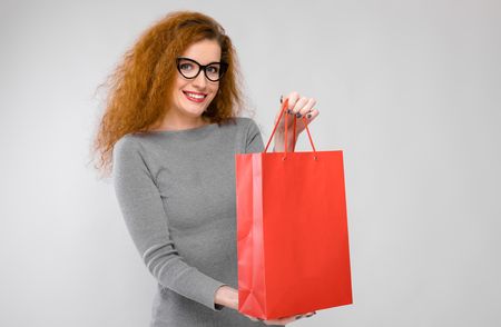 Portrait of attractive happy red-haired woman in dress and glasses isolated on grey background with copyspace holding colourful shopping paper bag.の写真素材