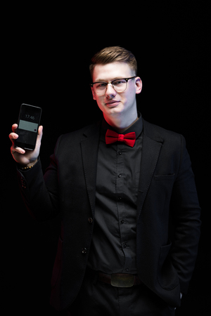 Young caucasian elegant successful smiling smart business man in black suit and red bow-tie standing in office looking in camera showing his mobile phoneの写真素材