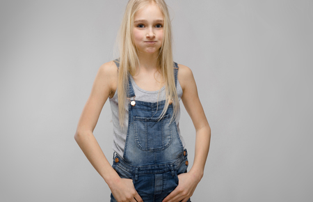 Portrait of beautiful teenage girl with blonde hair wearing denim overalls holding hands on pockets looking guilty isolated on grey background fashion advertising concept.の写真素材