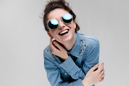 Young girl in round glasses on a gray background. Portrait of a young beautiful girl. Charming girl in a blue denim shirt.の写真素材