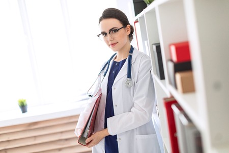 A charming young girl with glasses, a blue suit and a white coat is standing in a bright office. A stethoscope hangs around her neck. photo with depth of field.の写真素材