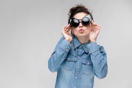 Young girl in black cat glasses on a gray background. Portrait of a young beautiful girl. Charming girl in a blue denim shirt.の写真素材