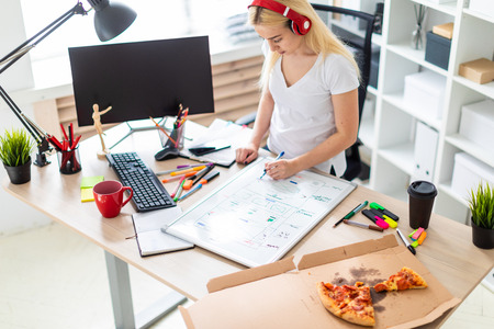 Beautiful blonde girl in a white tank top and dark pants. The girl listens to music through the red headphones and works with a marker and a magnetic board in a light cabinet. photo with depth of fieldの写真素材
