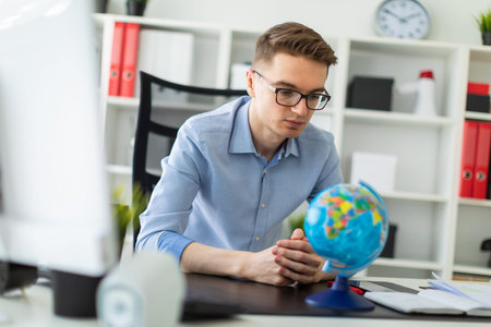 A handsome young man with glasses, blue trousers and a light shirt is working in the office. On the...