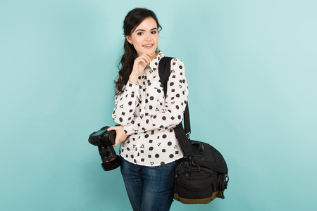 Portrait of young attractive woman photographer in white shirt holding camera and bag isolated on blue background with copyspace.の写真素材