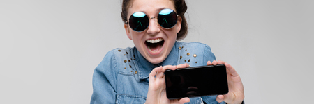 Young girl in round glasses on a gray background. Portrait of a young beautiful girl. Charming girl in a blue denim shirt. The girl is holding a phone.の写真素材