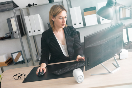 Beautiful young girl working in a bright office. Shes typing on the keyboard. The girl has blond hair. She is wearing a light blouse and a striped jacket. photo with depth of fieldの写真素材