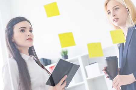 Two young beautiful girls are discussing something in a bright office. Girls are behind a transparent Board with stickers. They have business style clothes and beautiful long hair. photo with depth of fieldの写真素材