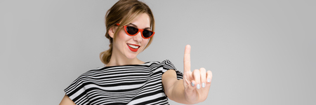 Portrait of woman wearing stylish sunglasses, striped top and pants holding her finger up in front of her isolated on grey background with copyspace fashion advertising or beauty salon concept.の写真素材