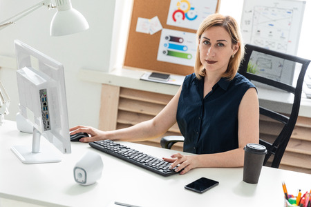 A beautiful young girl works in a bright office at a computer Desk. The girl has blond hair. She is wearing a light blouse and a striped suit. photo with depth of fieldの写真素材