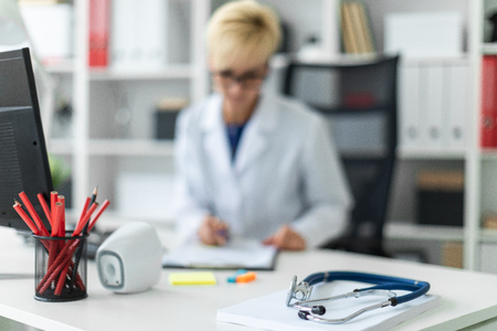 On the table is a phonendoscope. A beautiful young girl in a white robe working in a bright office at a computer Desk. She is holding a pen and a document. The girl has short white hair and wearing glasses. photo with depth of fieldの写真素材