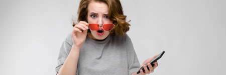 Beautiful young girl posing on gray background. A girl in red glasses is holding a phone in her hand