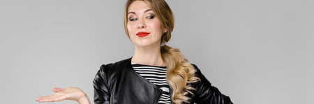 Portrait of woman wearing striped top, pants and leather jacket pointing with her palm at empty...
