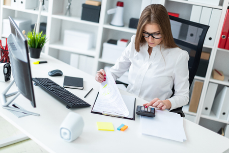Beautiful young girl working in a bright office. Young girl in white blouse and black trousers. She has long brown hair. Girl with glasses and beautiful makeup. Young girl sitting on a chair with a high back. photo with depth of fieldの写真素材