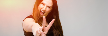 Half-length portrait of smiling young brunette caucasian girl in black dress on gray background showing peace signの写真素材