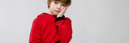 Portrait of upset little boy wearing red hoody holding hand on his cheek suffering from toothache isolated on grey background with copyspace.の写真素材