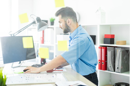 A smart young man works in a bright office. He has a neat beard. A young man in a blue shirt and dark jeans. photo with depth of fieldの写真素材
