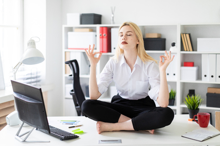 Cute young girl working in a bright office. She sits on the table in the Lotus position and relaxes. The girl has white hair and wearing glasses. Shes wearing a white shirt and black pants. photo with depth of fieldの写真素材