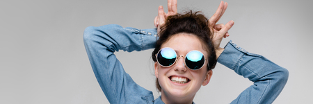 Young girl in round glasses on a gray background. Portrait of a young beautiful girl. Charming girl in a blue denim shirt.の写真素材