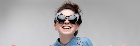 Young girl in black cat glasses on a gray background. Portrait of a young beautiful girl. Charming girl in a blue denim shirt. The girl has fun.の写真素材