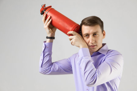 Portrait of a man in a shirt standing alone in a bright Studio and holding a red fire extinguisher near his ear. man checks the fire extinguisher. concept of fire safetyの写真素材