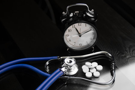 blue stethoscope and bunch of white pills lie on black background of a wooden table near clock....