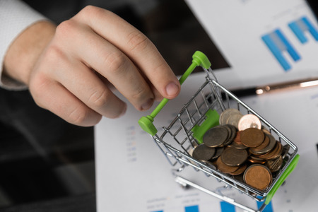 Hand holding a coin with pile of coin in the shopping cart on white and grey background. Symbolic photo for purchasing power and consumption. Finance and money concept mini shopping cart blurry backgroundの写真素材