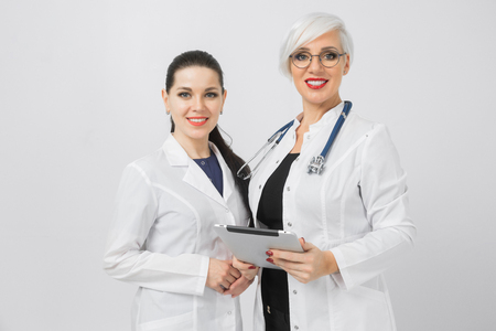 Two Young women in medical suits holding a digital tablet isolated on a light background. Horizontal portrait of Doctor and nurse standing in hospitalの写真素材